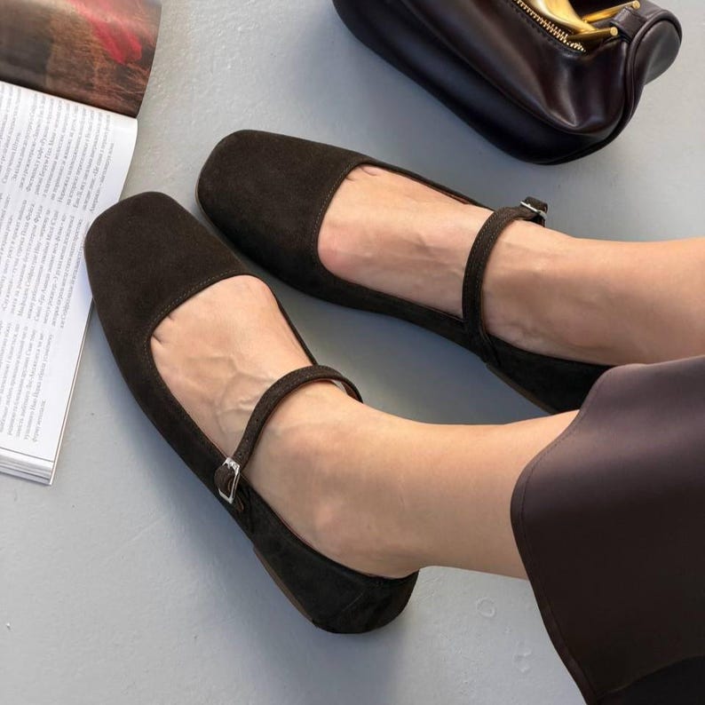 May include: A pair of dark brown suede flats with a square toe and ankle strap with a small buckle. The shoes are displayed on a light gray surface, with a dark brown purse in the background.