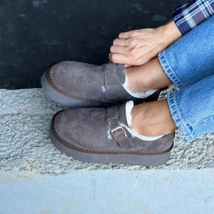 Könnte beinhalten: Graue Wildleder-Schlupfschuhe mit dicker, hellgrauer Sohle und Schnallen-Detail. Das Innere ist mit weißem Kunstfell gefüttert. Die Schuhe sind für Komfort und Wärme konzipiert, geeignet für den Alltag.