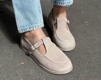 May include: A pair of light beige suede T-strap shoes featuring a rounded toe, a flat sole, and a thin adjustable strap with a silver-toned metal buckle across the top of the foot, worn with light blue denim jeans.