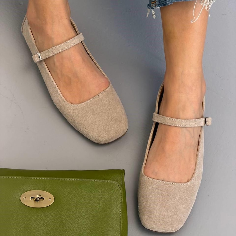 May include: Beige suede Mary Jane flats with a square toe and ankle strap with a small buckle. A green leather clutch with a gold clasp is also visible.