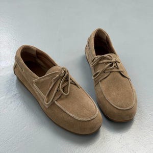 May include: A pair of tan suede boat shoes with light-colored laces. The shoes have a rounded toe and a stitched trim. The laces are tied in a bow. The shoes are displayed on a light gray surface.