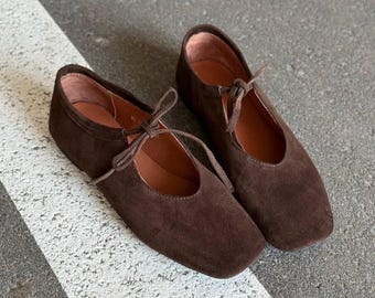 May include: A pair of brown suede ballet flats with a square toe and tie closure. The shoes have a low heel and a soft, textured appearance. The interior is a lighter brown color.