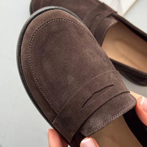 May include: A pair of dark brown suede loafers featuring a rounded toe, a classic strap across the vamp, and a textured black rubber sole. The shoes have a soft, matte finish and visible stitching along the perimeter of the toe box.
