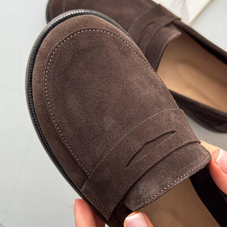 May include: A pair of dark brown suede loafers featuring a rounded toe, a classic strap across the vamp, and a textured black rubber sole. The shoes have a soft, matte finish and visible stitching along the perimeter of the toe box.