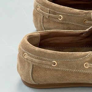 May include: Close-up of two tan suede boat shoes. The shoes have a classic design with a rounded toe, a leather sole, and gold-colored eyelets. The laces are tan and the interior is a darker brown.