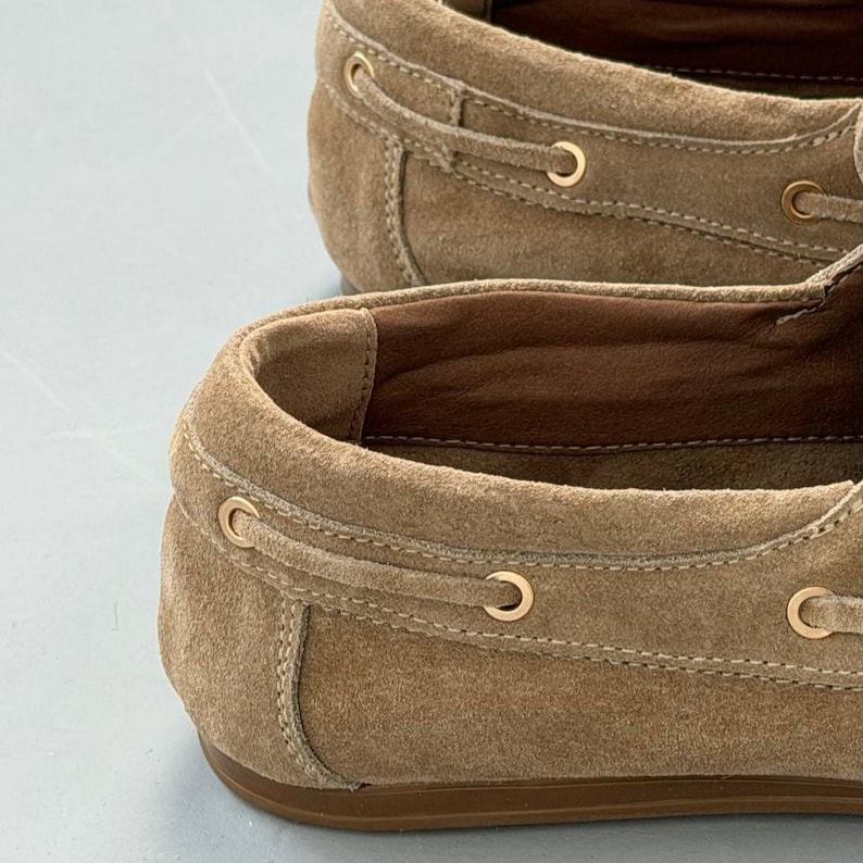 May include: Close-up of two tan suede boat shoes. The shoes have a classic design with a rounded toe, a leather sole, and gold-colored eyelets. The laces are tan and the interior is a darker brown.