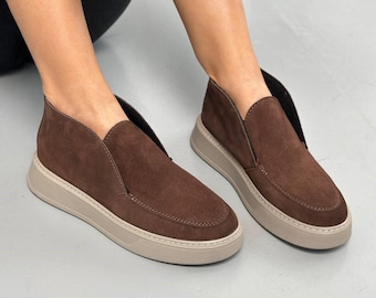 Op de afbeelding: Een paar donkerbruine suède instapschoenen met een laag uitgesneden enkelontwerp, een gestikte neus in mocassin-stijl en een dikke, getextureerde beige rubberen plateauzool. De schoenen hebben een strak silhouet zonder veters.