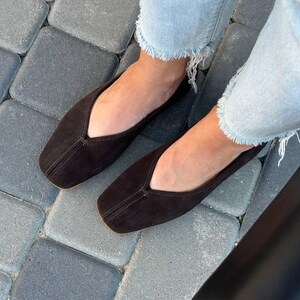 Pode incluir: Um par de sapatilhas de camur&ccedil;a castanhas escuras com bico quadrado. Os sapatos t&ecirc;m um decote baixo e costura central. Usadas com jeans azul claro. Cal&ccedil;ado elegante.