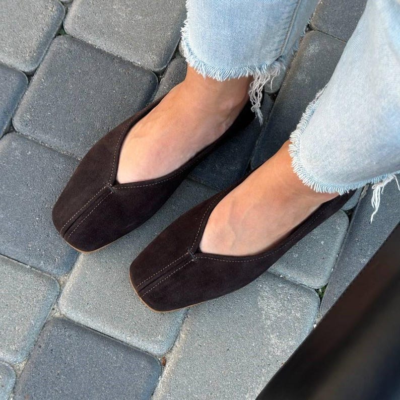 Pode incluir: Um par de sapatilhas de camur&ccedil;a castanhas escuras com bico quadrado. Os sapatos t&ecirc;m um decote baixo e costura central. Usadas com jeans azul claro. Cal&ccedil;ado elegante.