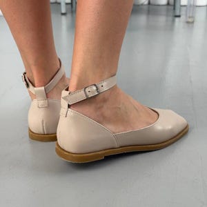 May include: Beige leather flats with ankle straps and small silver buckles. The shoes have a rounded toe and a flat sole. The image shows the shoes from a side view, highlighting the design and details of the footwear.