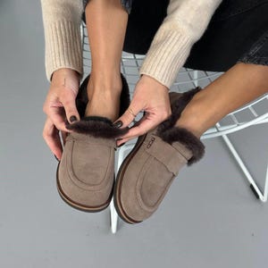 May include: A pair of taupe suede loafers featuring a dark brown faux fur lining and trim. The slip-on shoes have a rounded toe, a classic penny loafer strap across the vamp, and a dark brown sole with visible stitching along the edge of the base.
