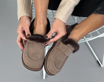 Könnte beinhalten: Ein Paar taupefarbene Wildleder-Loafer mit dunkelbraunem Kunstfellfutter und Besatz. Die Slipper haben eine abgerundete Zehenpartie, einen klassischen Penny-Loafer-Riemen über dem Vorderblatt und eine dunkelbraune Sohle mit sichtbaren Nähten.