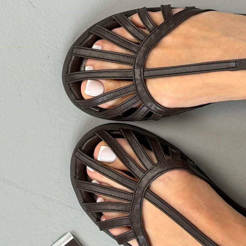Puede incluir: Primer plano de dos sandalias de cuero marr&oacute;n con un dise&ntilde;o similar a una jaula. Las sandalias tienen m&uacute;ltiples correas que crean un patr&oacute;n tejido en la parte superior del pie. Las sandalias est&aacute;n sobre una superficie gris.