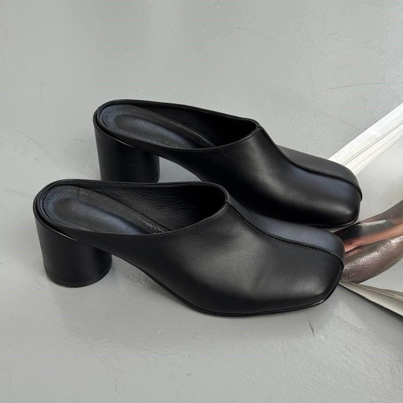 May include: A pair of black leather mules with a square toe and a block heel. The heels are approximately 3 inches high. The shoes are displayed on a light gray surface.