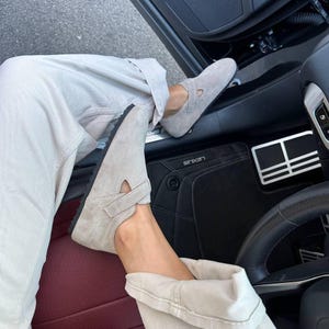 May include: A pair of light gray suede shoes with a strap closure. The shoes have a rounded toe and a black sole. The shoes are worn with light-colored pants.