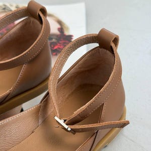 May include: Close-up of brown leather sandals with ankle straps and a small buckle closure. The sandals have a rounded toe and a low heel. The straps are stitched for durability.