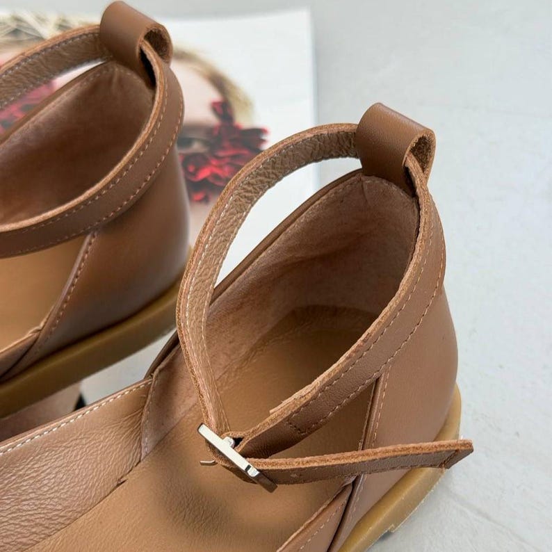May include: Close-up of brown leather sandals with ankle straps and a small buckle closure. The sandals have a rounded toe and a low heel. The straps are stitched for durability.