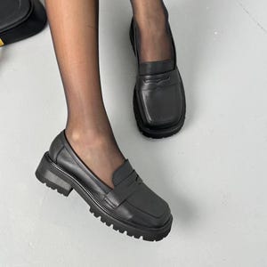 May include: A pair of black leather loafers featuring a square toe design, a classic penny slot strap across the vamp, and a thick, chunky lug sole with deep treads. The shoes are worn with sheer black hosiery, showcasing the smooth texture.