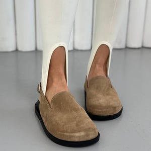 Könnte beinhalten: Ein Paar beigefarbene Wildleder-Slipper mit abgerundeter Zehenpartie, einem Fersenriemen mit kleiner Metallschnalle und einer dicken schwarzen Gummisohle. Die Schuhe werden mit weißen Steghosen getragen, die eine Aussparung am Fersenbereich haben.