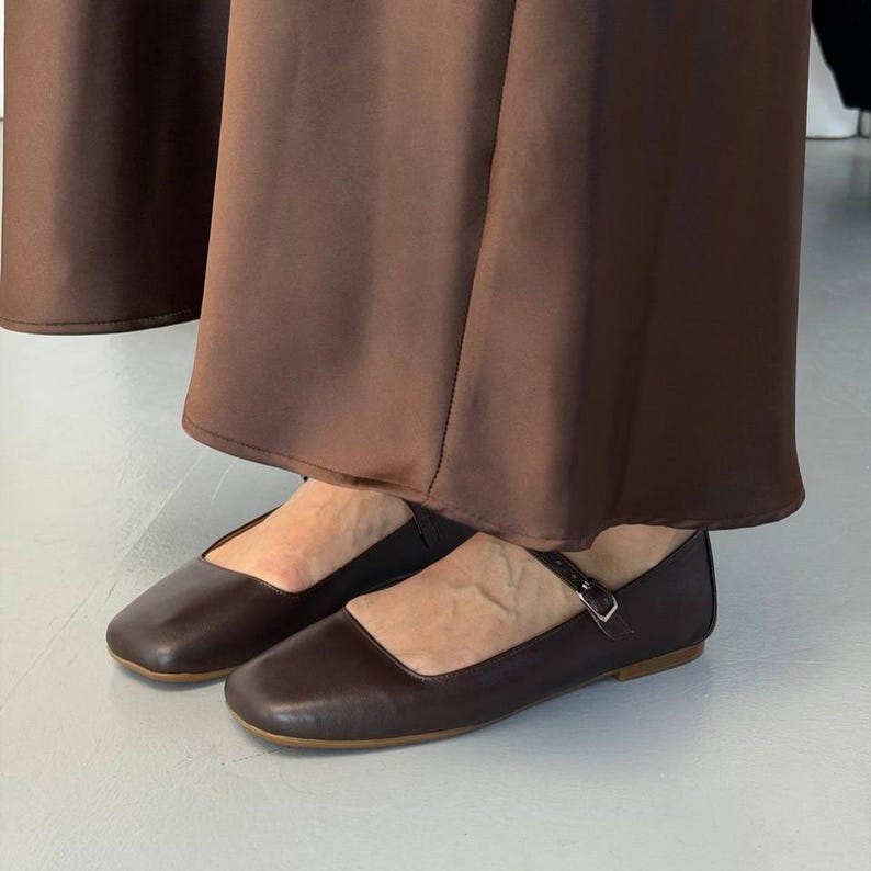 May include: A pair of dark brown Mary Jane flats with a square toe and a small buckle closure. The shoes are worn with a long, flowing brown skirt.