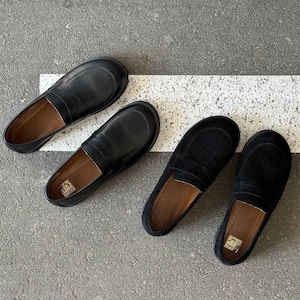May include: Four pairs of black leather and suede penny loafers are arranged on a gray surface with a white stripe. Two pairs are smooth leather, and two are suede. The shoes have a classic design with a strap across the vamp.