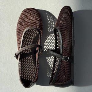 May include: A pair of dark brown Mary Jane flats featuring a breathable mesh upper with a hexagonal pattern. Each shoe includes a thin, adjustable strap across the instep with a small silver-toned metal buckle and solid trim along the edges.