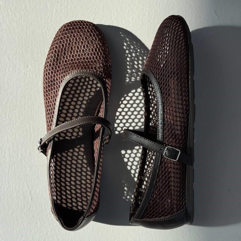 May include: A pair of dark brown Mary Jane flats featuring a breathable mesh upper with a hexagonal pattern. Each shoe includes a thin, adjustable strap across the instep with a small silver-toned metal buckle and solid trim along the edges.