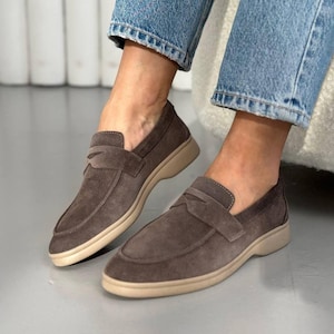 May include: A pair of dark taupe suede loafers featuring a classic penny strap across the vamp, a rounded toe, and a thick, flat beige rubber sole. The shoes are worn with blue denim jeans, showcasing a low-cut silhouette and tonal stitching.