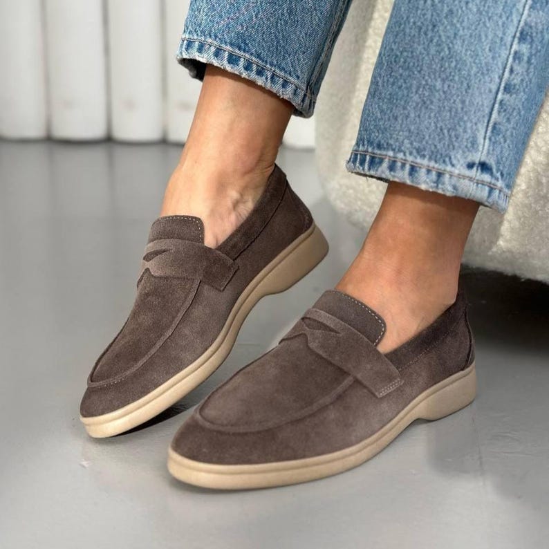 May include: A pair of dark taupe suede loafers featuring a classic penny strap across the vamp, a rounded toe, and a thick, flat beige rubber sole. The shoes are worn with blue denim jeans, showcasing a low-cut silhouette and tonal stitching.