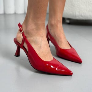 May include: A pair of glossy red slingback heels with pointed toes. The shoes have a small, curved heel and a delicate strap that fastens with a gold buckle. The shoes are a vibrant shade of red.