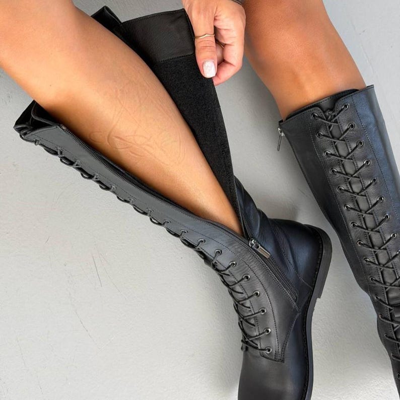 May include: A pair of black knee-high boots featuring a lace-up front design with black laces and metal eyelets. The boots include a full-length side zip closure that extends from the sole to the top, revealing a black interior lining.