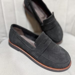 May include: A pair of black suede loafers featuring a classic penny slot design across the vamp. The shoes have a rounded toe, a low profile, and a dark sole with a visible brown stitched welt detail running along the edge of the base.