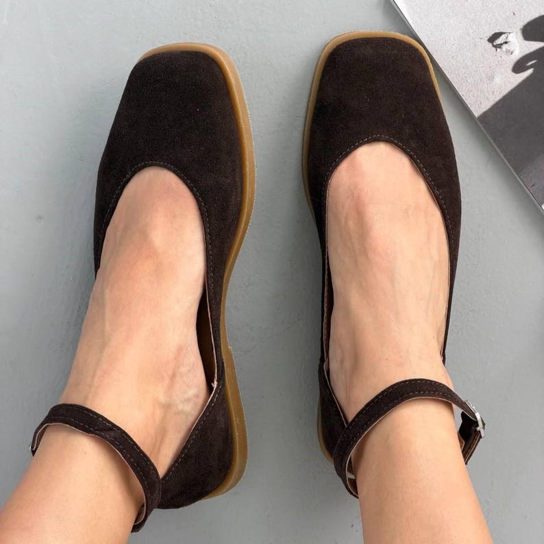May include: A pair of dark brown suede flats with a square toe and ankle strap. The shoes have a light brown sole and a buckle closure on the ankle strap. The flats are designed for comfort and style.