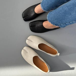 May include: Black and off-white leather split-toe flats. The black flats are worn by a person wearing blue jeans. The off-white flats are displayed on a gray surface. The shoes have a unique design with a split between the big toe and other toes.