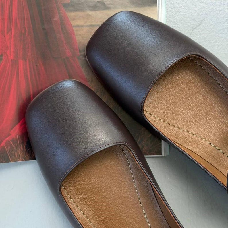 May include: Close-up of two brown leather flats with square toes. The shoes have a smooth, matte finish and a light brown interior. The stitching is visible along the edges of the shoes.