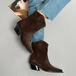 Peut inclure: Une paire de bottes de cowboy en daim marron avec un bout pointu et un talon épais. Les bottes ont un design western classique avec des coutures décoratives et une tirette. Les bottes sont portées avec un jean bleu.