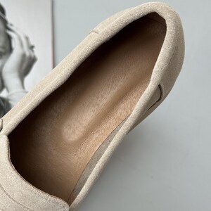May include: A beige suede slip-on shoe featuring a soft, textured exterior and a smooth, tan-colored interior lining. The shoe has a rounded toe design and a clean, minimalist construction with visible stitching along the edges of the upper material.