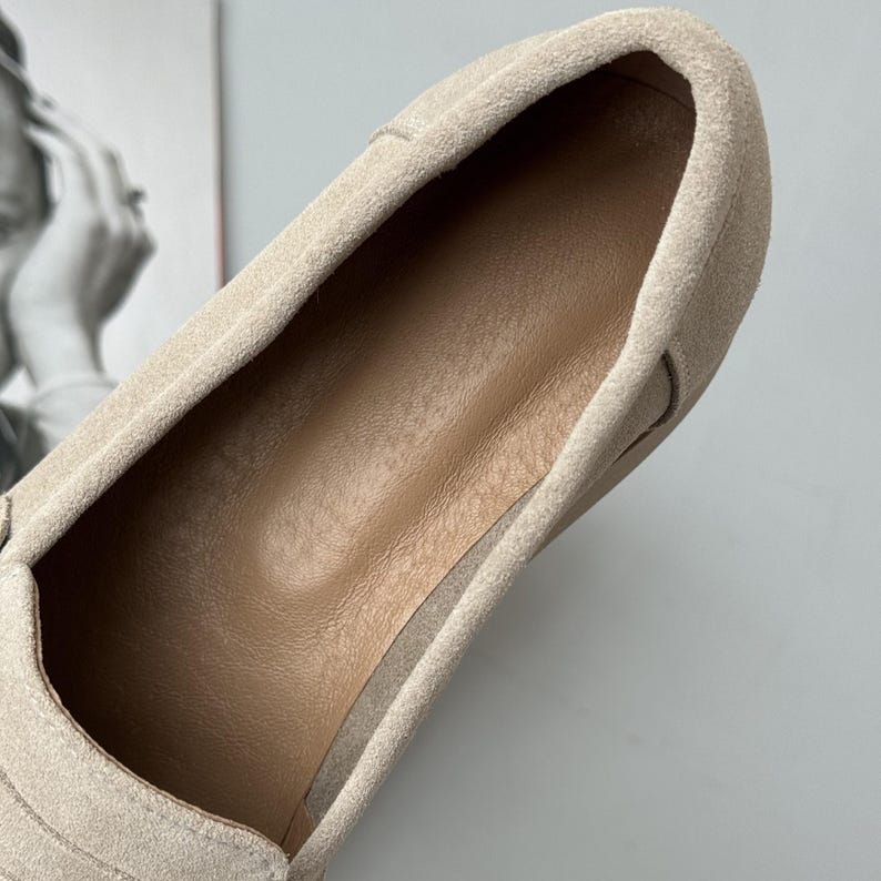 May include: A beige suede slip-on shoe featuring a soft, textured exterior and a smooth, tan-colored interior lining. The shoe has a rounded toe design and a clean, minimalist construction with visible stitching along the edges of the upper material.