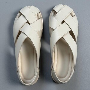 May include: A pair of off-white, woven leather sandals. The sandals feature a crisscross design across the top, with a rounded toe and a flat sole. The shoes are displayed on a gray surface.