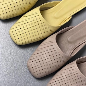 May include: A pair of slip-on mules featuring a square toe and a perforated upper design. One shoe is a bright pastel yellow, while the other is a neutral beige. Both shoes have a smooth interior lining and a low-profile, flat construction.