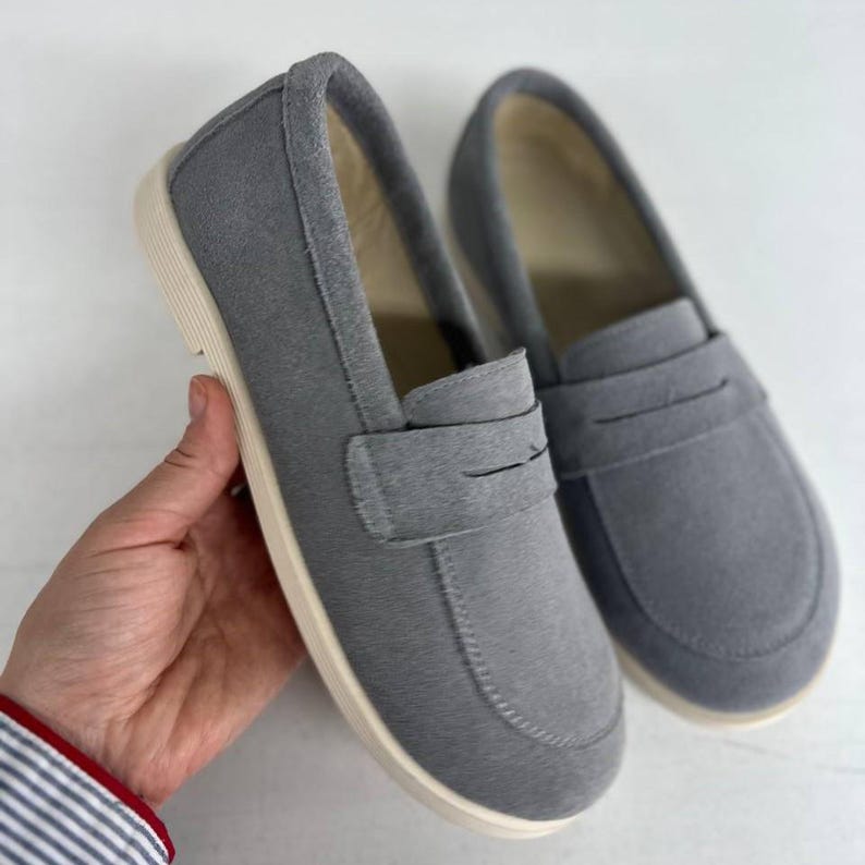 May include: A pair of gray suede loafers with a classic penny strap design. The shoes have a rounded toe and a cream-colored sole. These casual slip-on shoes are suitable for everyday wear.
