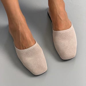 May include: A pair of beige slip-on mules featuring a square toe design and a perforated texture across the upper surface. These flat shoes have a simple, open-back construction that allows for easy wear, showcasing a consistent light neutral color.