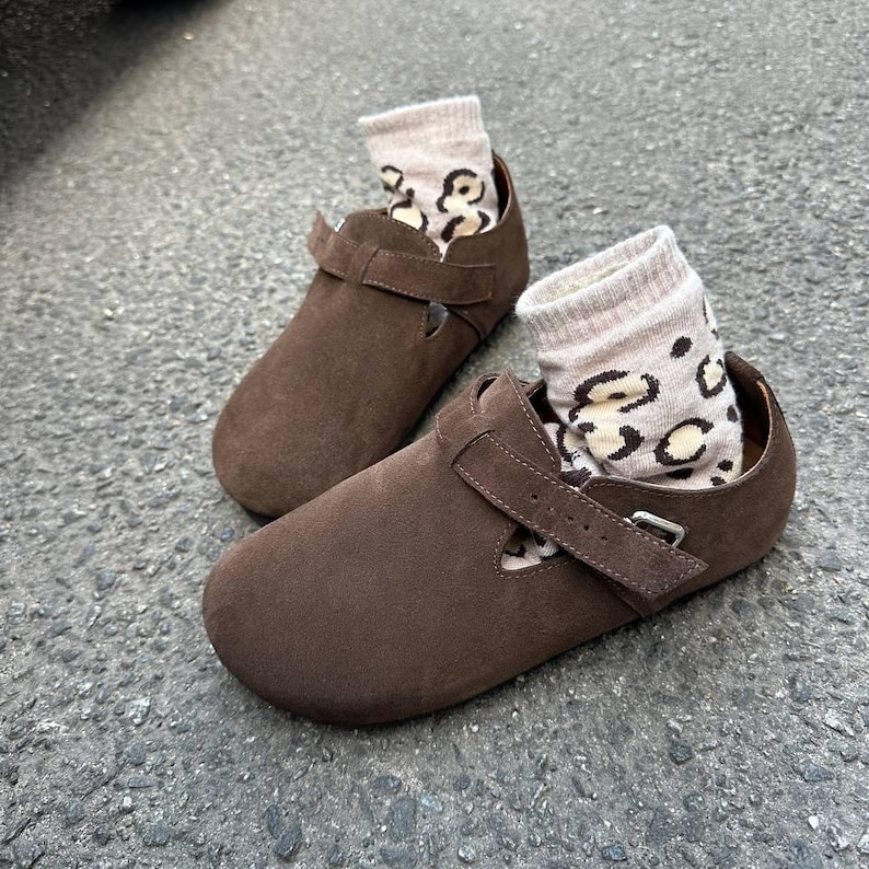 May include: A pair of brown suede Mary Jane style shoes with a strap and buckle closure. The shoes are filled with beige socks featuring a black leopard print. The shoes are sitting on a gray surface.