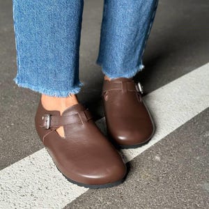 May include: Brown leather shoes with a buckle closure. The shoes have a rounded toe and a strap across the top. The shoes are worn with blue jeans.