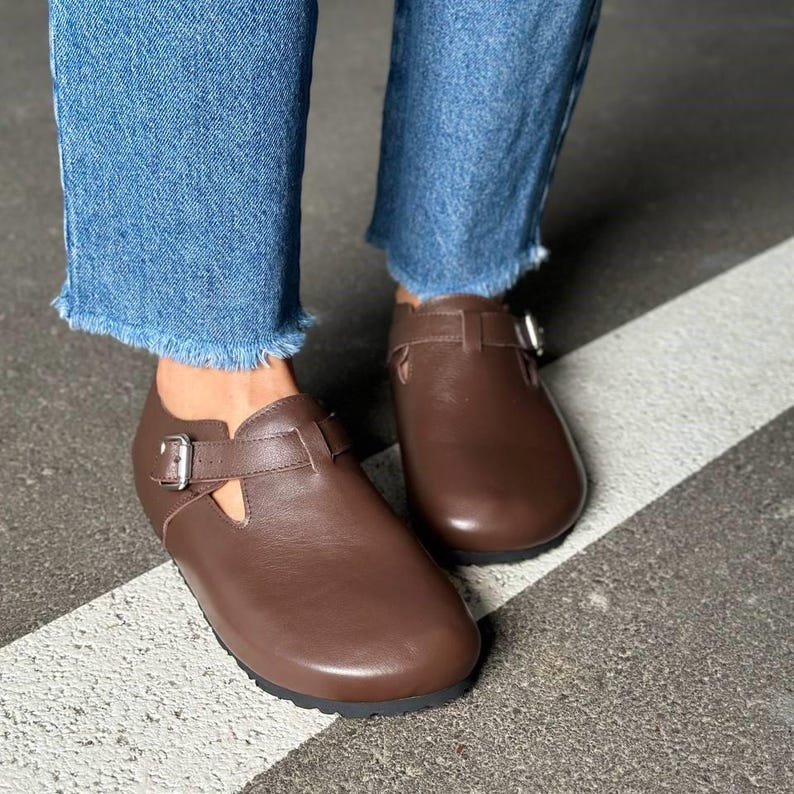 May include: Brown leather shoes with a buckle closure. The shoes have a rounded toe and a strap across the top. The shoes are worn with blue jeans.
