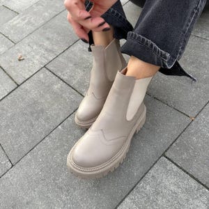 May include: A pair of beige ankle boots featuring a smooth leather-like finish, elastic side panels, a rounded toe with decorative wingtip stitching, and a thick, lugged rubber sole. The boots are worn with dark gray denim jeans.