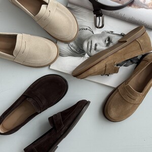 May include: A collection of suede loafers in beige, tan, and dark brown colors. Each shoe features a classic penny loafer strap across the vamp, a rounded toe, and a flat sole. The shoes are arranged in a group, showcasing their soft texture.