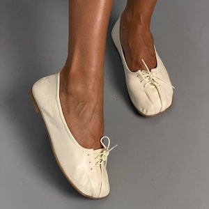 Op de afbeelding: Een paar off-white ballerina's met een zachte, ronde neus en een centraal vetersluitingsdetail met dunne, bijpassende koordjes die in een kleine strik aan de bovenkant zijn geknoopt. De schoenen hebben een laag profiel en een gladde afwerking.