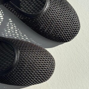 May include: A pair of black mesh flats featuring a rounded toe design and a solid black trim along the opening. The upper material consists of a fine, breathable grid-like mesh fabric that creates a textured appearance across the surface of the shoes.