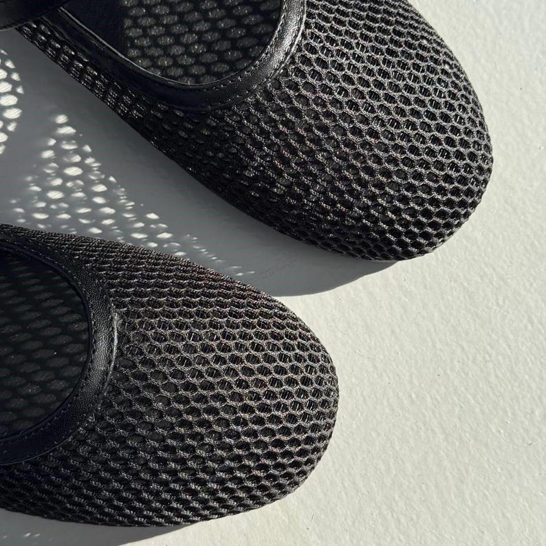 May include: A pair of black mesh flats featuring a rounded toe design and a solid black trim along the opening. The upper material consists of a fine, breathable grid-like mesh fabric that creates a textured appearance across the surface of the shoes.